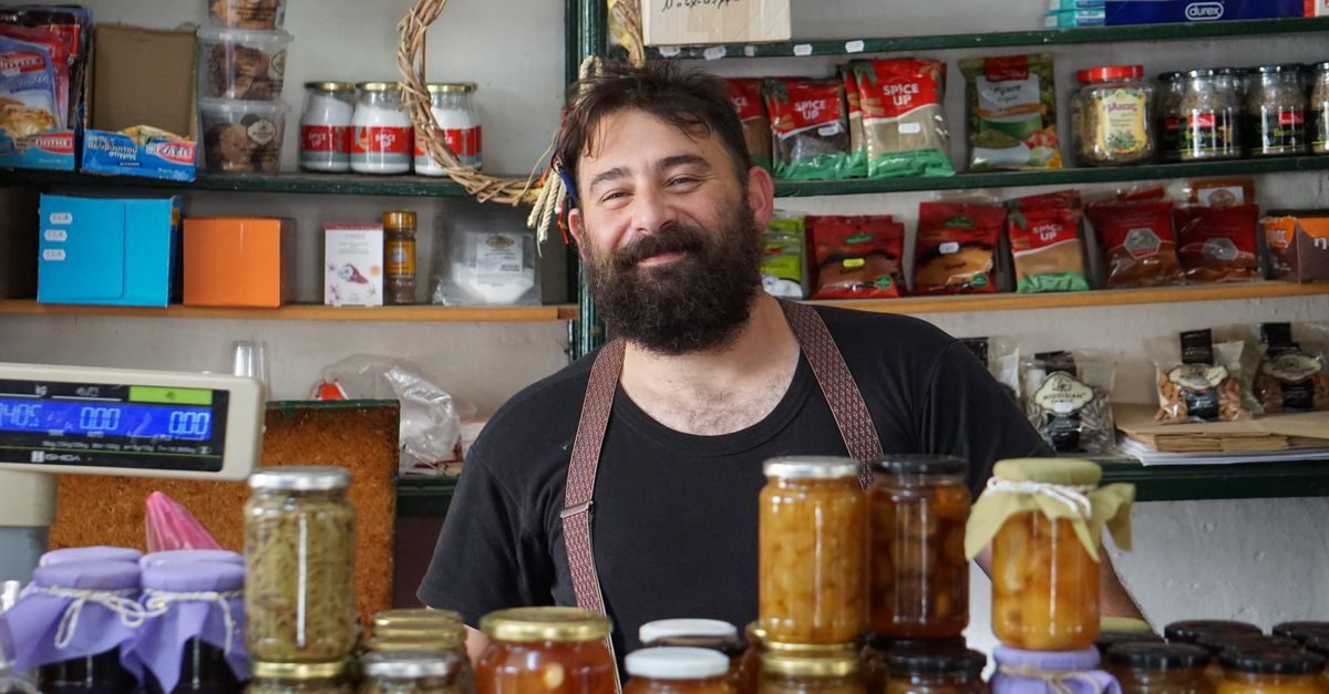 Hombre con barba y tirantes sonríe detrás del mostrador de una tienda de comestibles con estantes llenos de frascos y bolsas