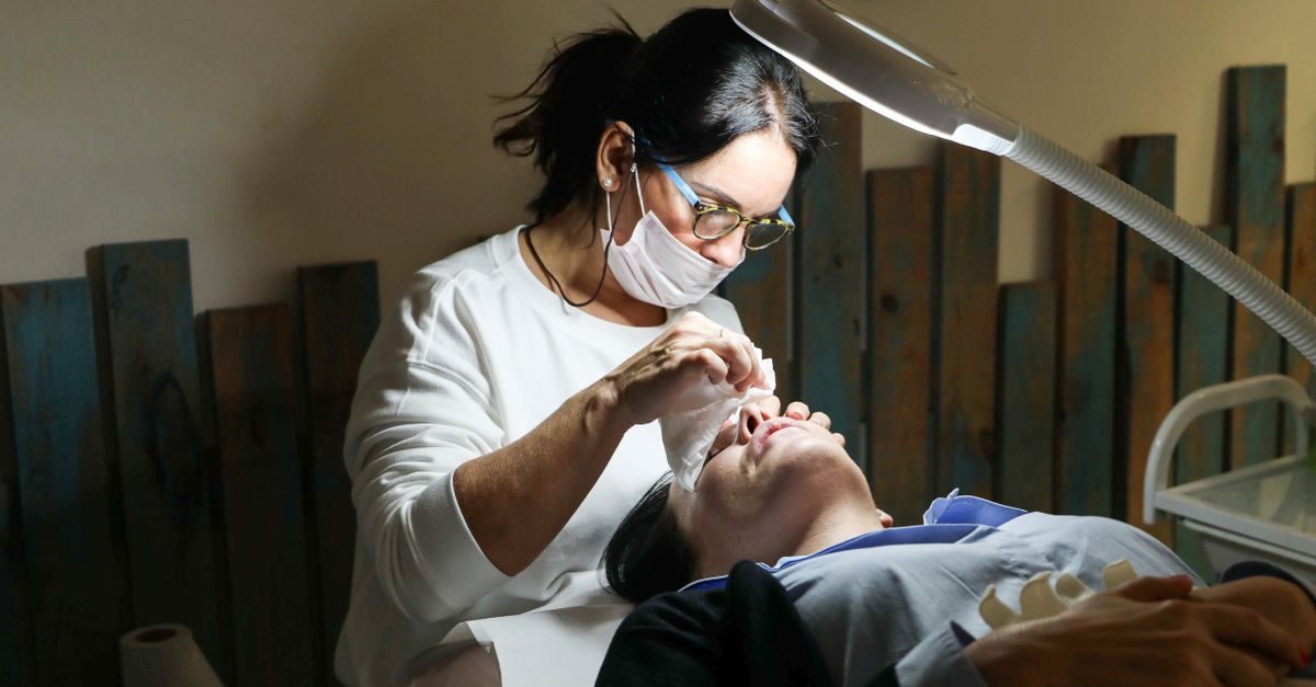 Kosmetikerin mit Mundschutz und Brille behandelt unter hellem Licht das Gesicht einer liegenden Kundin im Studio