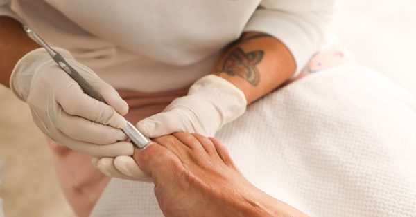 Persona con guantes de látex realiza una limpieza de cutícula en el pie de un cliente con un instrumento metálico