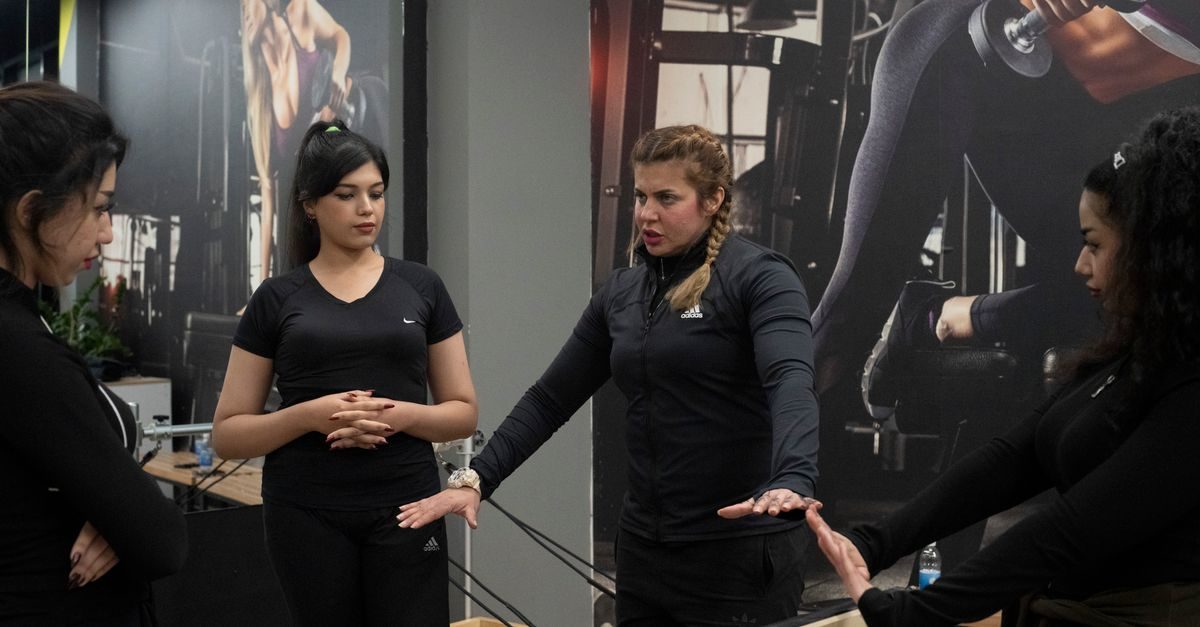 Fitnessinstructrice geeft uitleg aan drie vrouwen in een sportschool met grote muurposters op de achtergrond