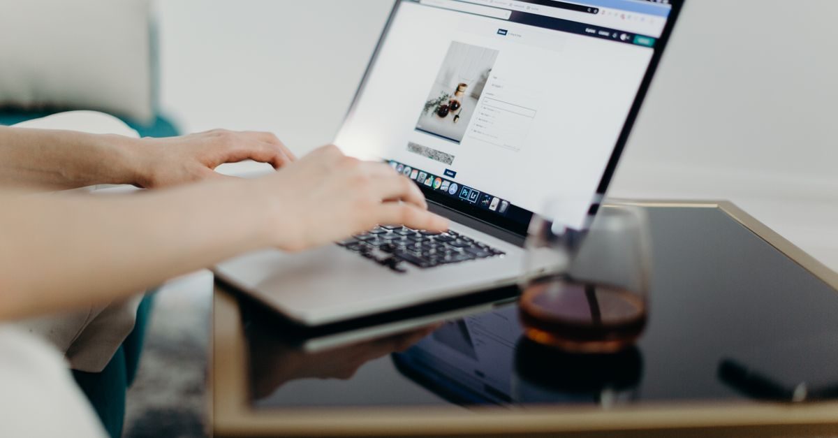 Hände tippen auf einer Laptop-Tastatur an einem Tisch mit einem Glas Getränk in einer hellen, modernen Arbeitsumgebung