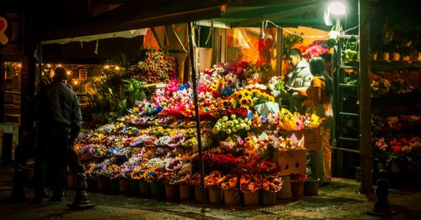 Puesto de flores nocturno con clientes y dependientes bajo una iluminación cálida en una calle urbana