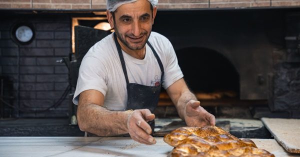 Panadero sonriente con gorro y delantal coloca pan recién horneado sobre una mesa frente a un horno de piedra tradicional