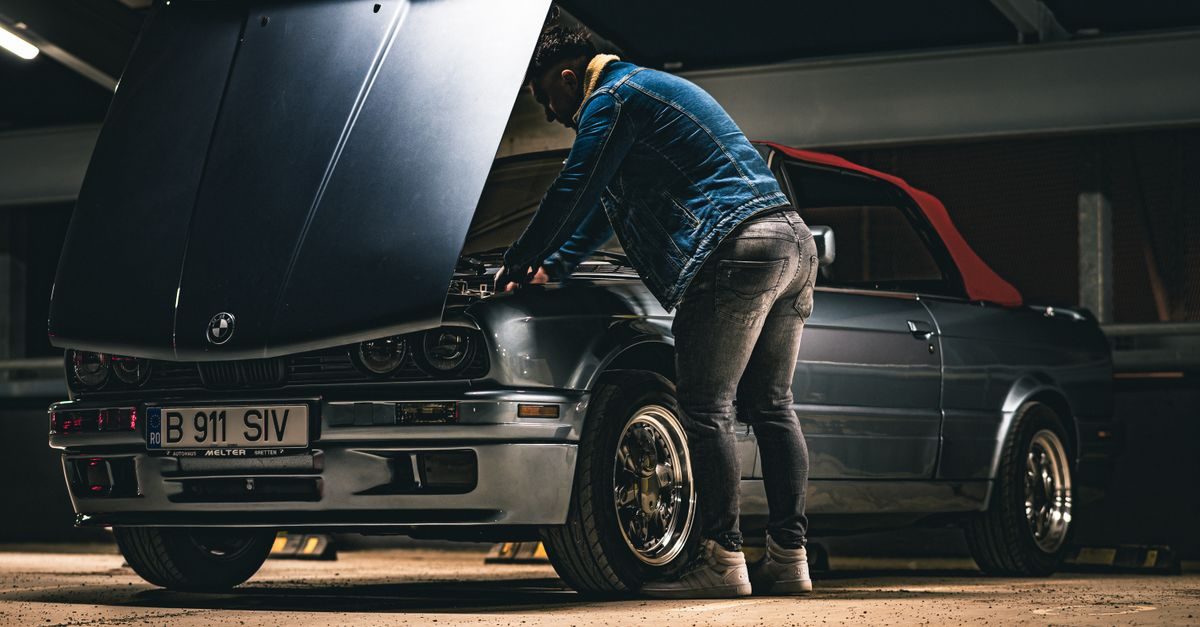 Un ragazzo in giacca di jeans controlla il motore di una BMW cabrio d'epoca in un garage sotterraneo con luci soffuse