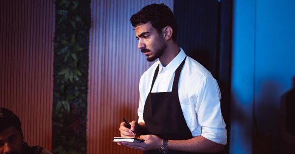 Camarero con camisa blanca y delantal negro toma nota concentrado en una libreta dentro de un restaurante con luz tenue