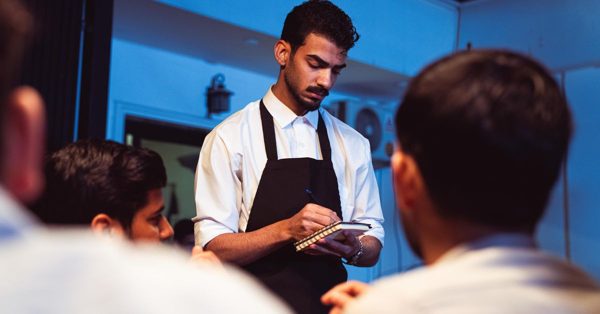 A focused waiter in a white shirt and black apron takes an order from customers in a dimly lit cafe