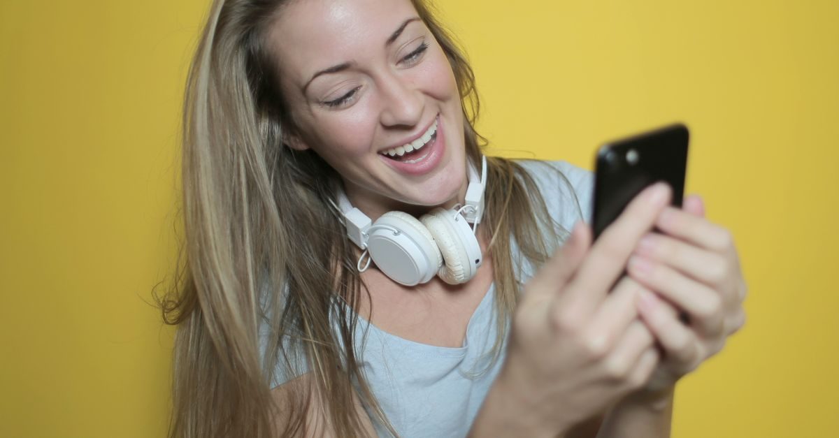 Uśmiechnięta blondynka ze słuchawkami na szyi patrzy z radością w ekran smartfona na żółtym tle