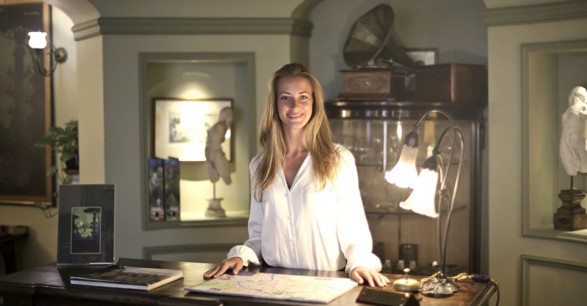 A smiling woman in a white shirt stands behind a reception desk in a warmly lit, classically decorated hotel lobby