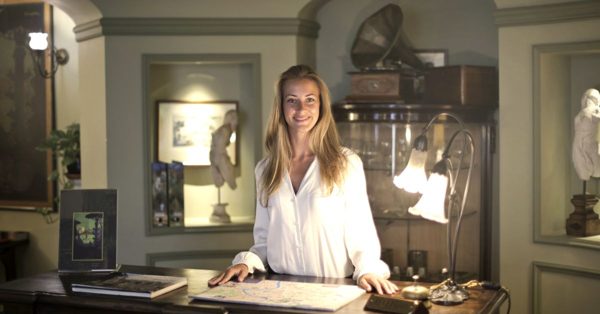 A smiling woman in a white shirt stands behind a reception desk in a warmly lit, classically decorated hotel lobby