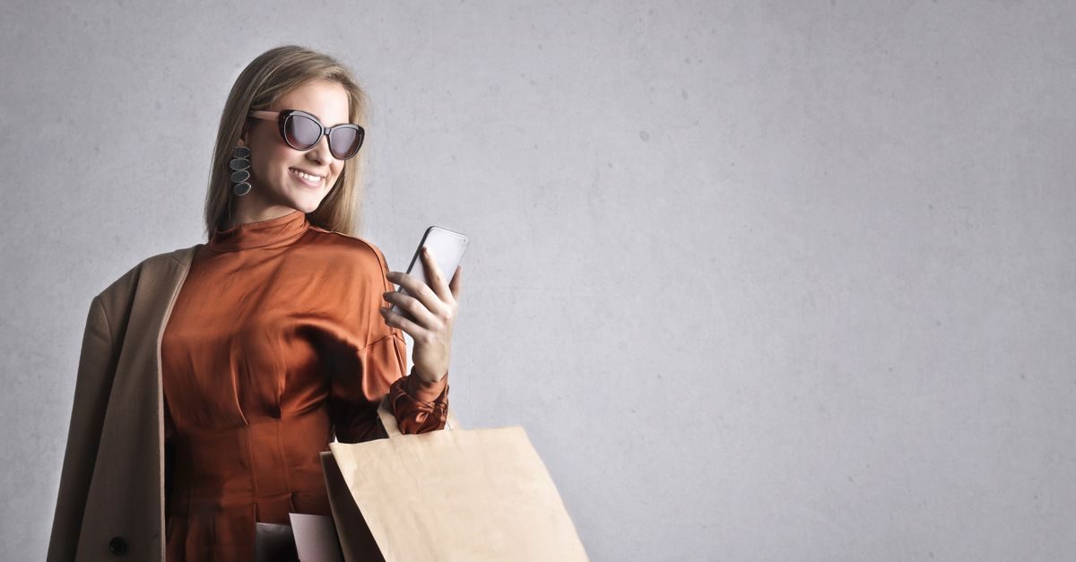 Mujer sonriente con gafas de sol y bolsas de compras mira su teléfono móvil frente a una pared gris clara