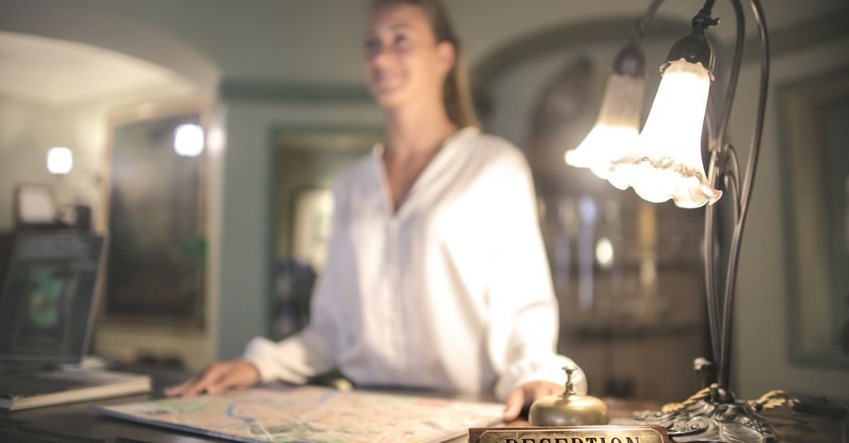 Eine freundliche Hotelmitarbeiterin steht hinter einer Rezeption mit einer Tischlampe und einer Glocke im Vordergrund