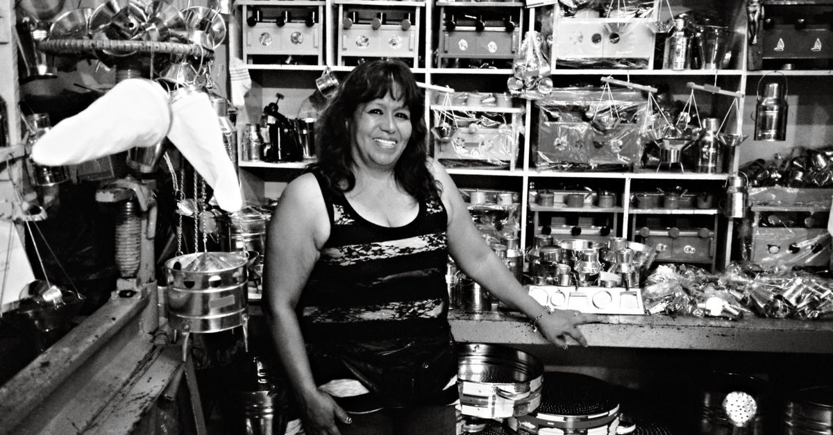 Une femme souriante se tient debout dans sa boutique remplie d'ustensiles et d'objets métalliques en noir et blanc