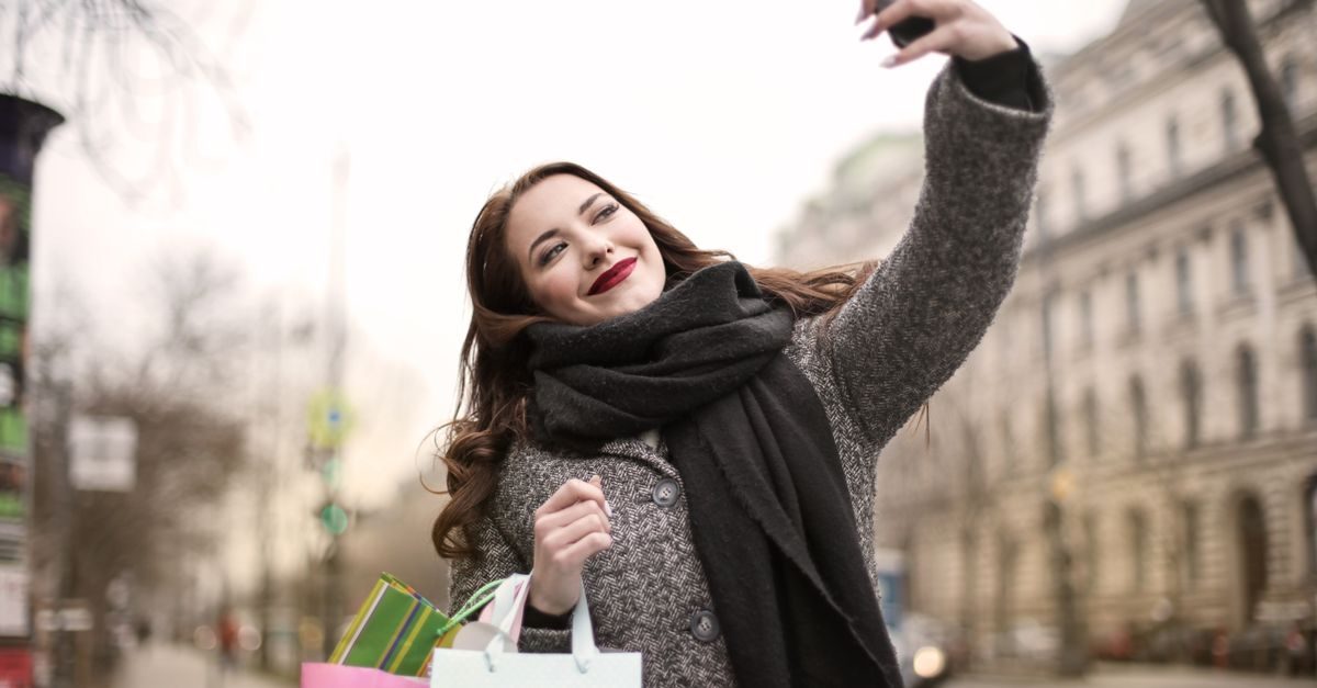 Eine junge Frau mit Schal und Einkaufstüten macht in einer städtischen Umgebung fröhlich ein Selfie mit ihrem Smartphone