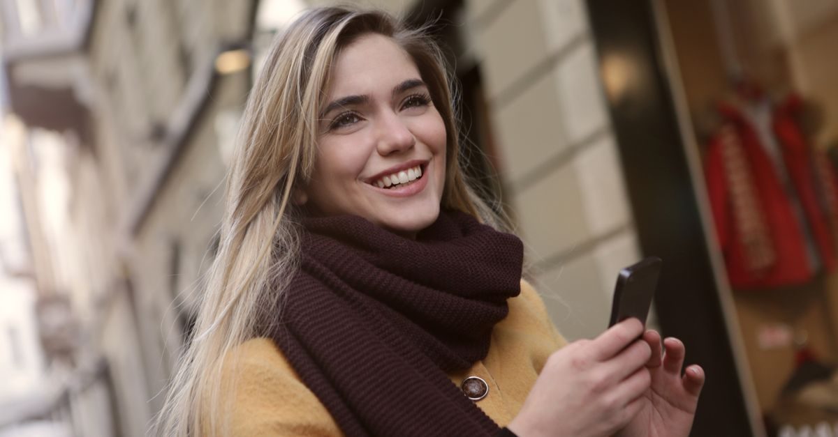 Mujer joven rubia sonriente con bufanda marrón y abrigo amarillo sostiene un teléfono móvil en una calle urbana