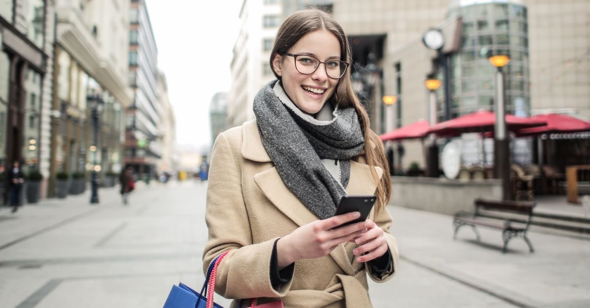 A smiling woman in a beige coat and scarf holds a smartphone while walking on a city street with shopping bags
