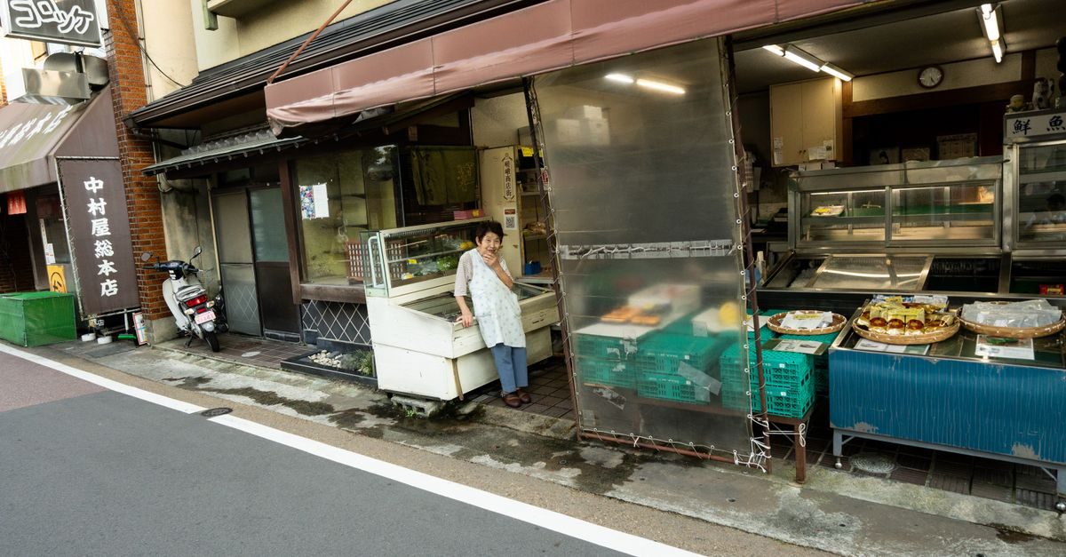 Vrouw in schort staat voor een kleine Japanse winkel met uitgestalde koopwaar aan de straatkant