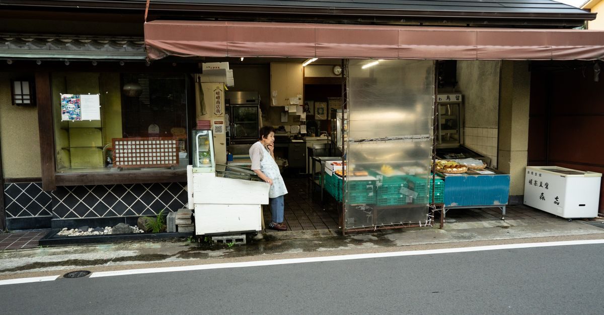 Een oudere vrouw staat in de deuropening van een traditionele Japanse winkel met etenswaren aan een rustige straat