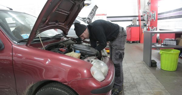 Mekaniker i svart mössa och arbetskläder inspekterar motorn på en röd bil inne i en bilverkstad
