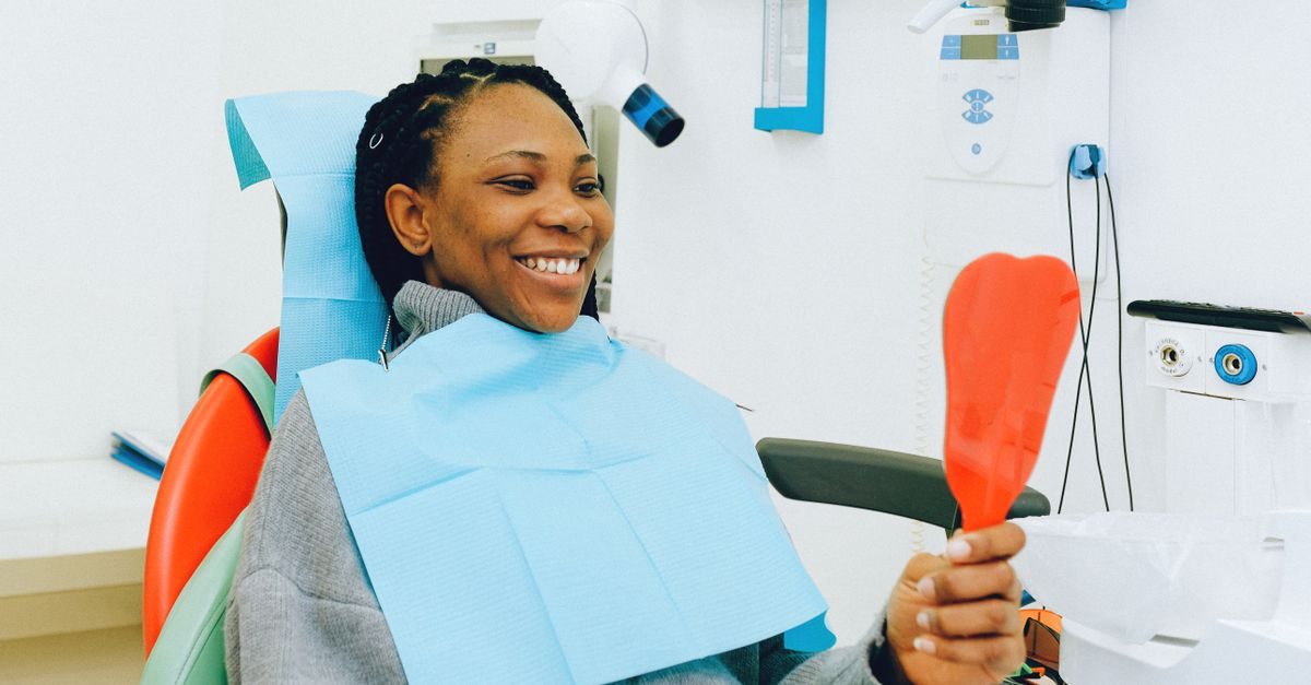 A smiling woman sits in a dental chair wearing a blue bib and holding a red mirror to check her teeth in a clinic