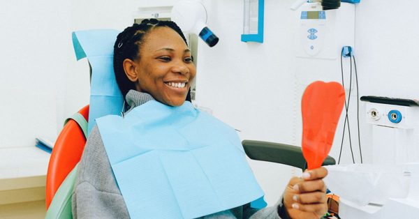 A smiling woman sits in a dental chair wearing a blue bib and holding a red mirror to check her teeth in a clinic
