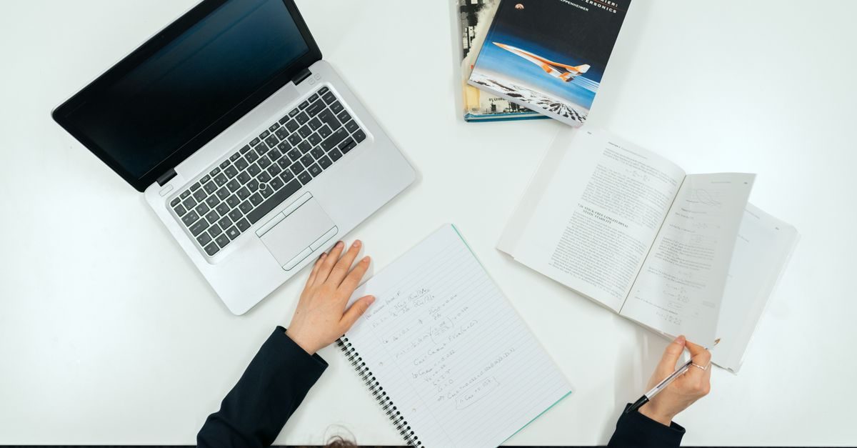 Vista cenital de una persona trabajando con un portátil, un cuaderno y libros sobre una mesa blanca de oficina