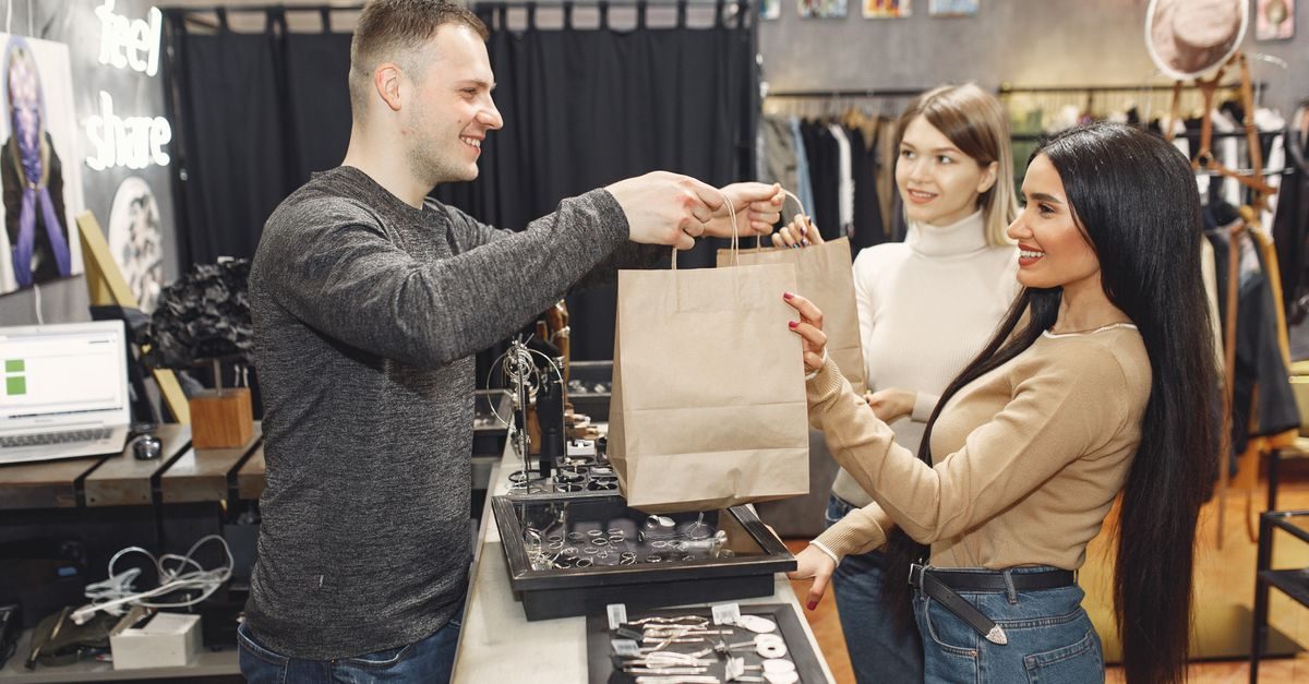 Ein freundlicher Verkäufer überreicht zwei lächelnden Kundinnen in einem Bekleidungsgeschäft ihre Einkaufstüten