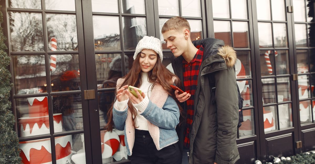 Giovane coppia sorridente guarda lo smartphone davanti a una vetrina decorata per le festività natalizie