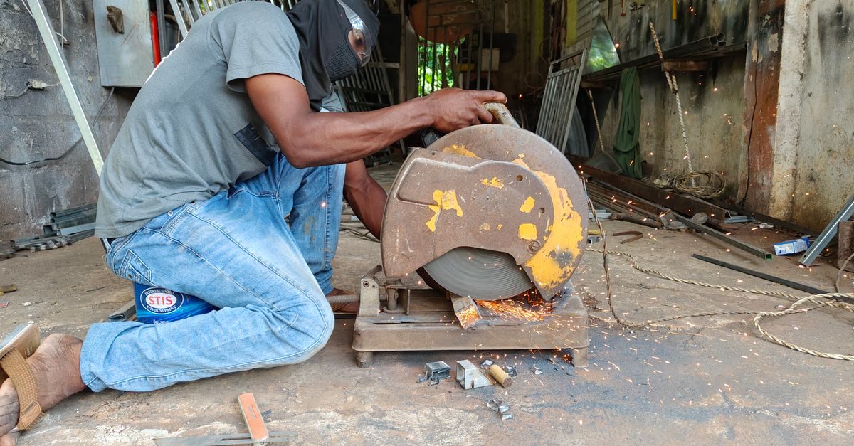 A worker wearing a face covering uses a metal chop saw to cut steel in a cluttered workshop, sending sparks flying