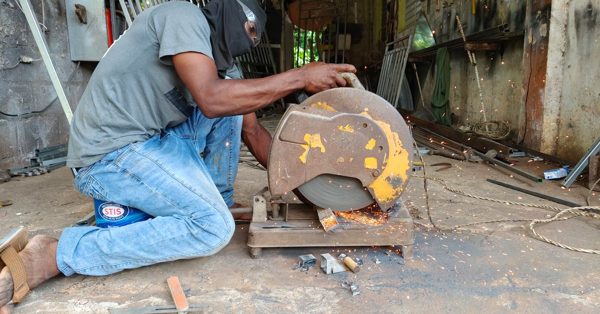 A worker wearing a face covering uses a metal chop saw to cut steel in a cluttered workshop, sending sparks flying