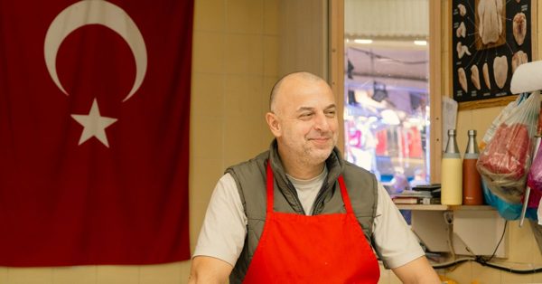 Un boucher souriant porte un tablier rouge devant un drapeau turc dans sa boutique