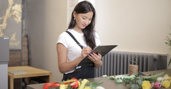 Une jeune femme souriante écrit sur un presse-papier dans un atelier de fleuriste avec des fleurs sur le comptoir
