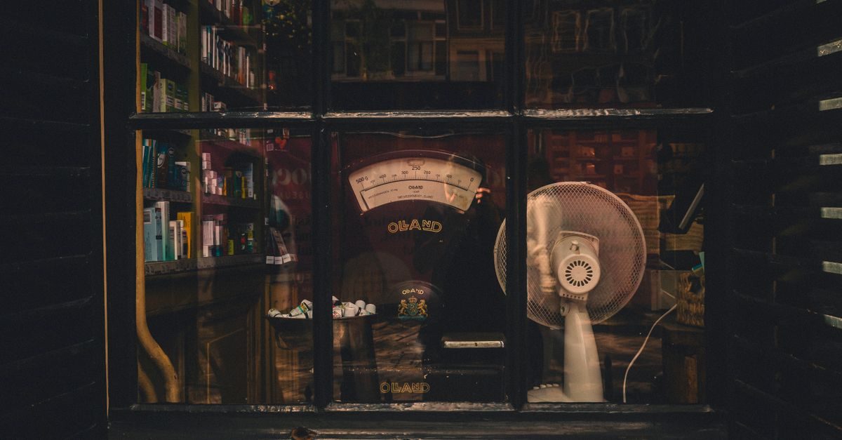 Donker winkelraam met een oude weegschaal, een ventilator en planken vol boeken in een sfeervolle, nostalgische setting