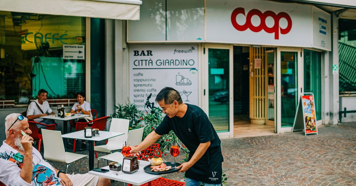 Ober serveert drankjes en hapjes op een terras voor een Italiaanse bar en een Coop-supermarkt