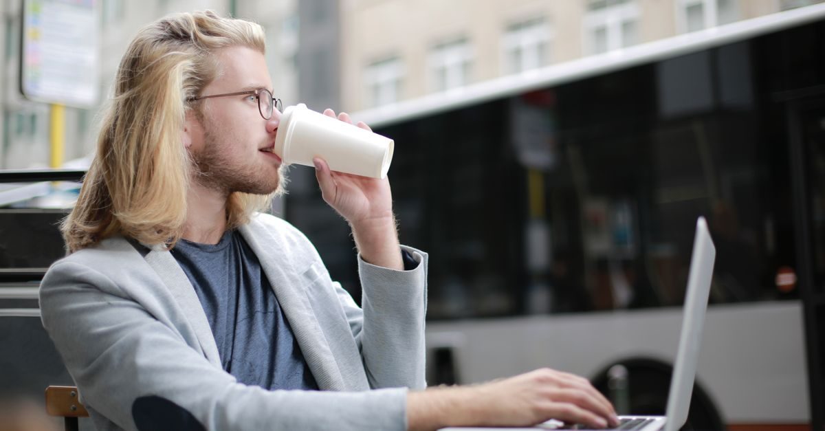 Jonge man met bril en blond haar drinkt koffie uit een beker terwijl hij op een laptop werkt op een terras in de stad