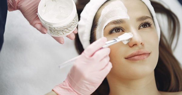Schoonheidsspecialist brengt met een kwastje een wit gezichtsmasker aan bij een ontspannen klant in een studio