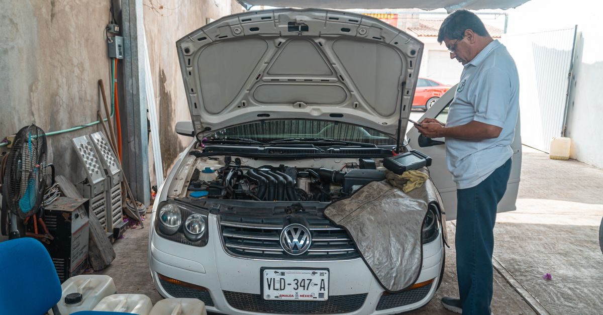 Ein Mechaniker prüft in einer Werkstatt mit geöffneter Motorhaube an einem weißen VW-Pkw die Fahrzeugdaten auf seinem Handy