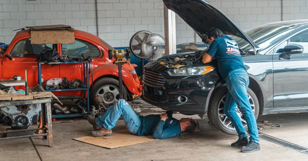 Zwei Mechaniker reparieren in einer Werkstatt einen schwarzen Pkw, während daneben ein roter Oldtimer steht