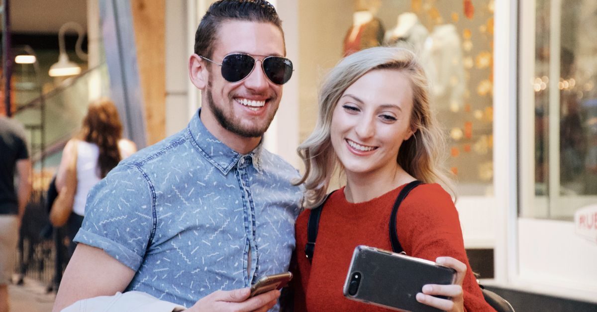 A smiling man and woman take a selfie together outside a storefront in a cheerful, casual setting