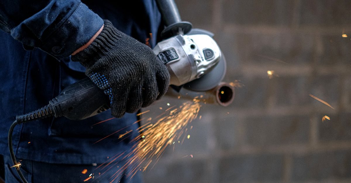 A worker in a blue uniform and black gloves uses an angle grinder to cut a metal pipe, creating a spray of bright sparks