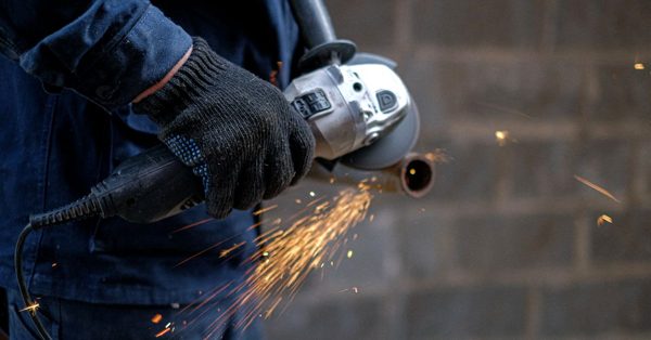 A worker in a blue uniform and black gloves uses an angle grinder to cut a metal pipe, creating a spray of bright sparks