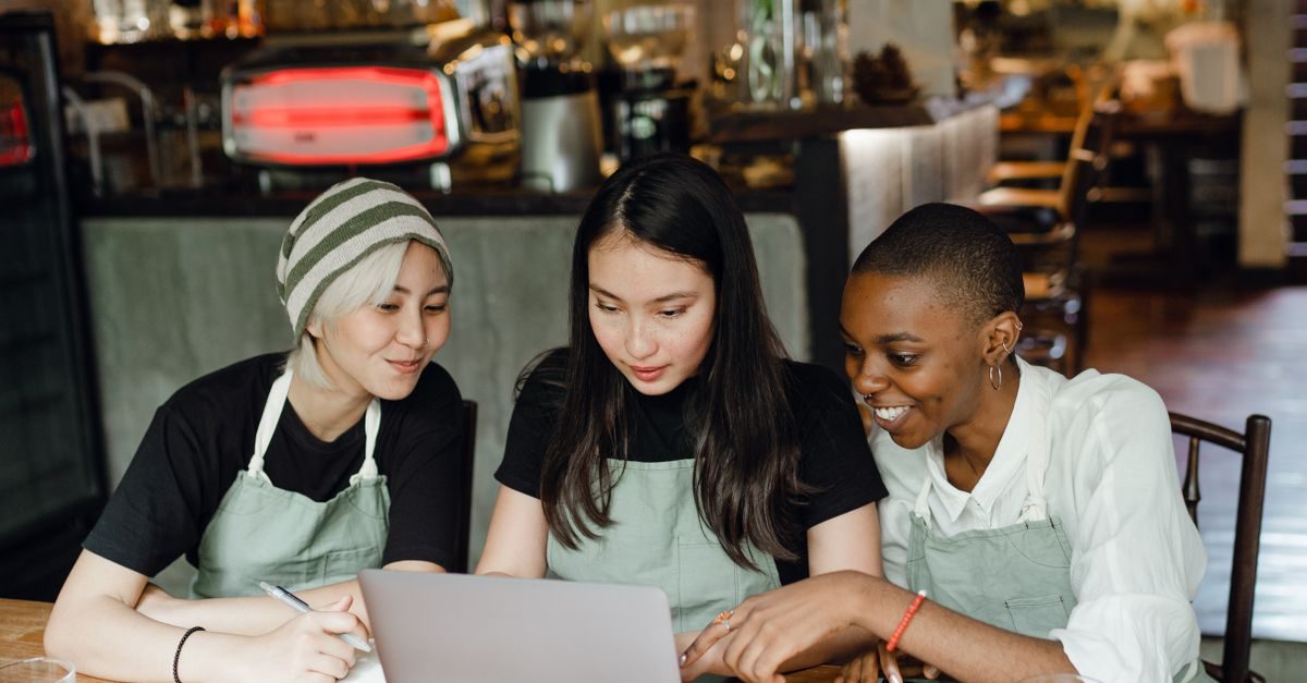 Trois employées en tablier travaillent ensemble sur un ordinateur portable dans un café à l'ambiance conviviale