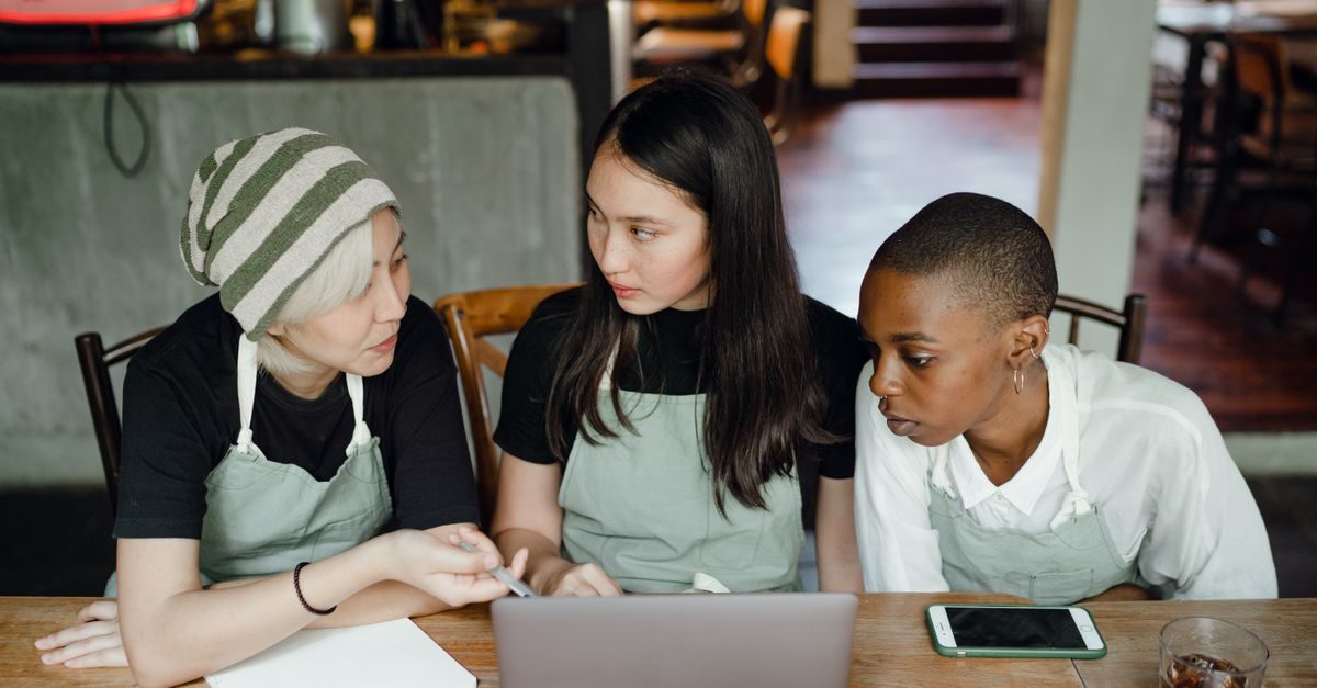 Three young women wearing aprons sit at a wooden table, focused on a laptop screen in a cafe setting