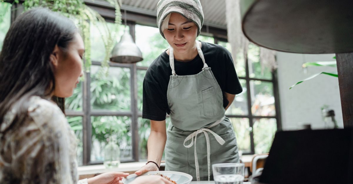 Vriendelijke ober met schort serveert een bord eten aan een klant in een licht restaurant met veel planten