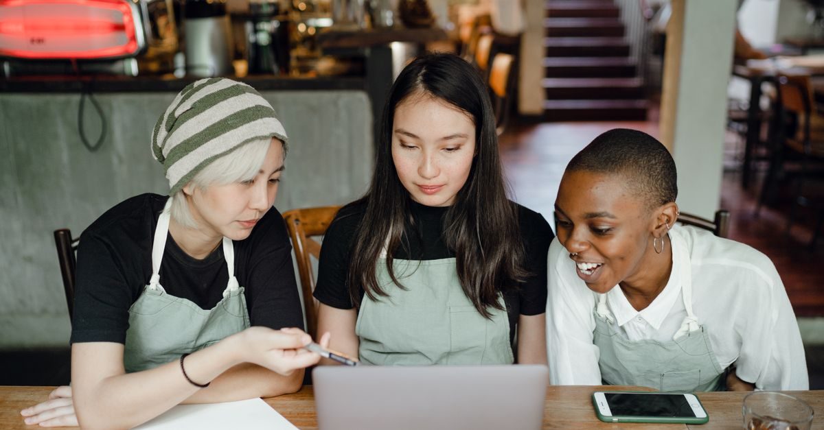 Drei junge Frauen in Arbeitskleidung sitzen in einem Café und schauen konzentriert und lächelnd auf einen Laptop