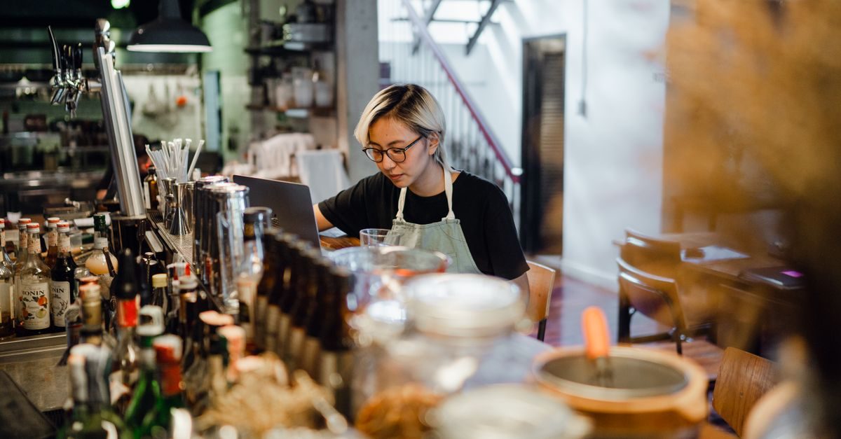 Geconcentreerde horecamedewerker met bril werkt achter een bar op een laptop tussen flessen drank in een rustig café