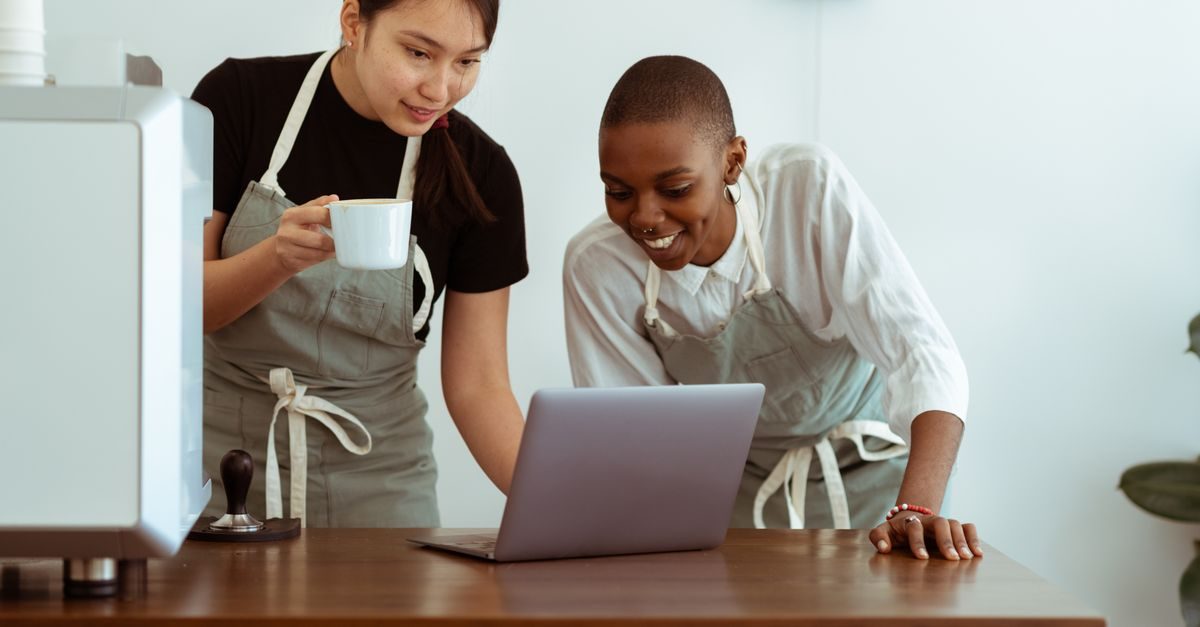 Dos empleadas de cafetería con delantales miran sonrientes una computadora portátil sobre una mesa de madera