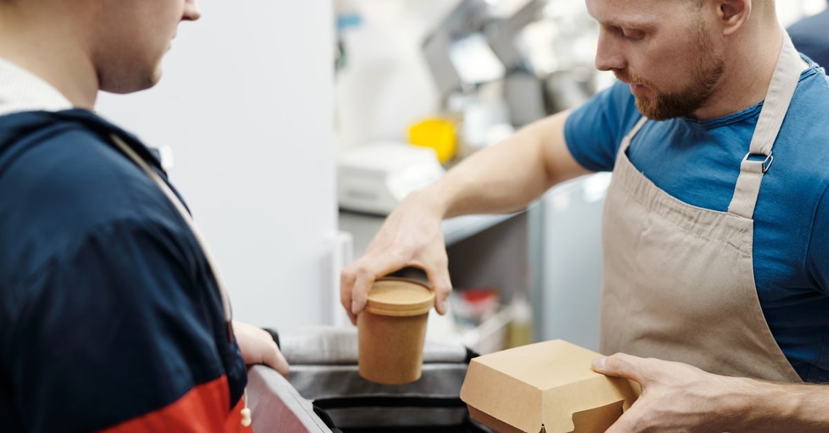 A focused cafe worker in an apron packs food into a delivery bag for a customer in a professional kitchen setting