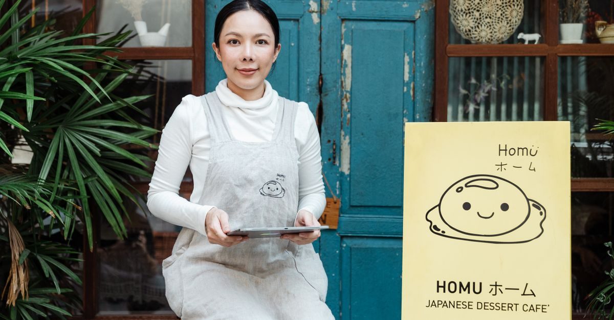 Une femme souriante en tablier tient une tablette devant l'entrée d'un café japonais avec une enseigne jaune à l'extérieur