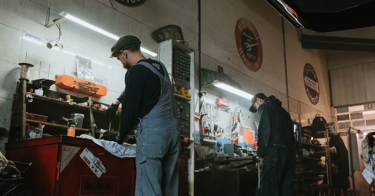 Twee monteurs in werkkleding werken geconcentreerd aan voertuigonderdelen in een sfeervolle, klassieke garage