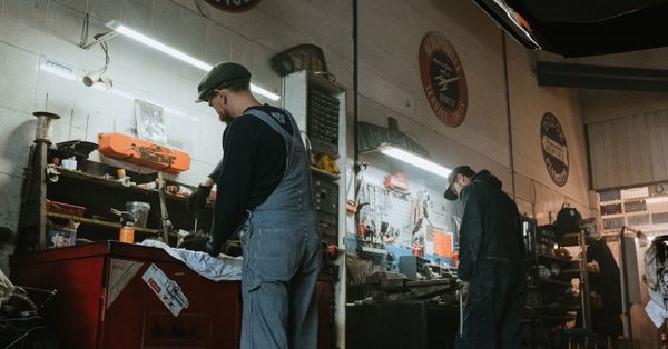 Twee monteurs in werkkleding werken geconcentreerd aan voertuigonderdelen in een sfeervolle, klassieke garage